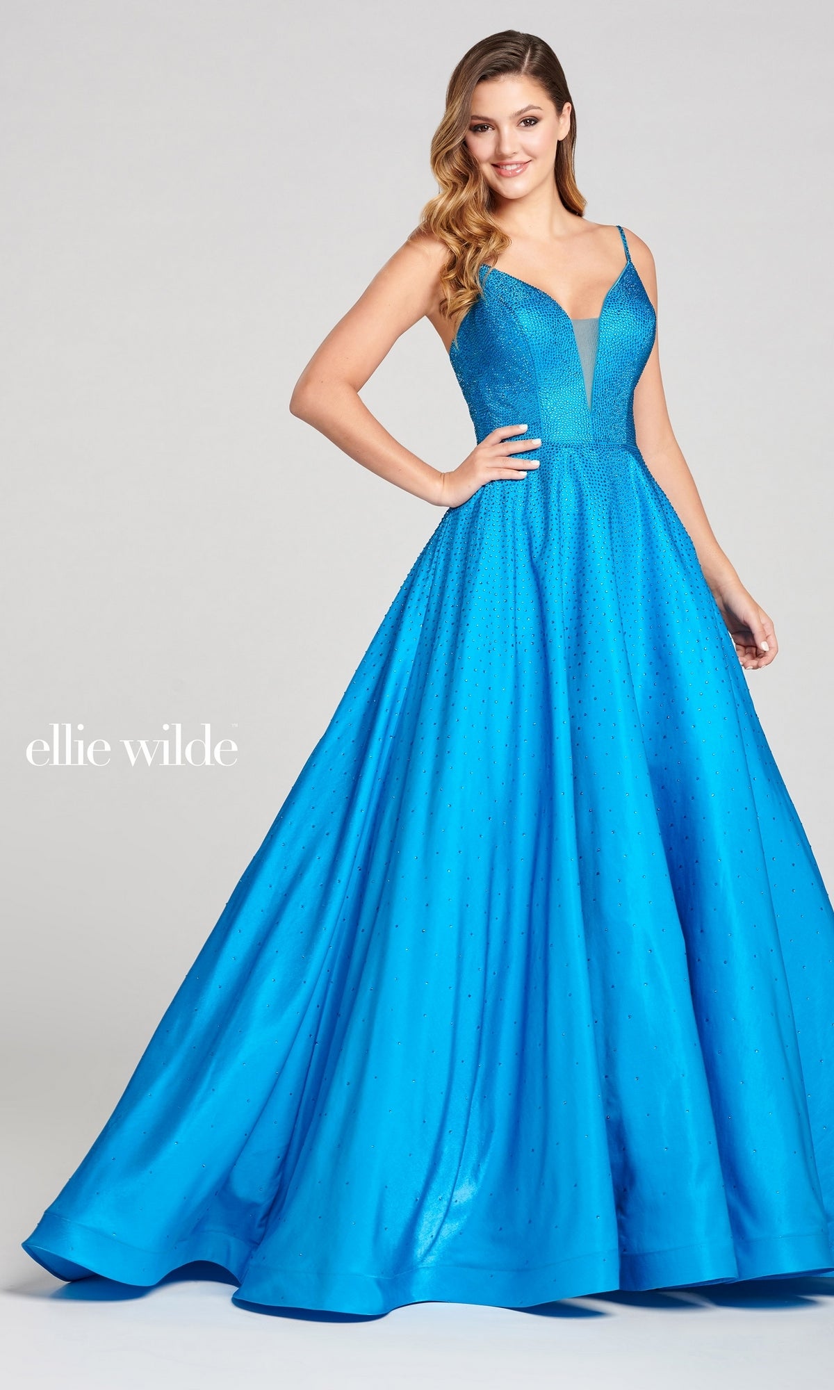 A woman poses in a bright blue, sleeveless Long Beaded Prom Ball Gown (Ellie Wilde Ew121005) with a fitted bodice and full skirt. The background is plain and